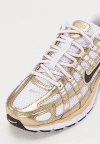 Białe i złote sneakersy Nike wykonane z siateczkowego materiału, z teksturowaną powierzchnią, misternym wzorem sznurowadeł i czarnym logo Swoosh.