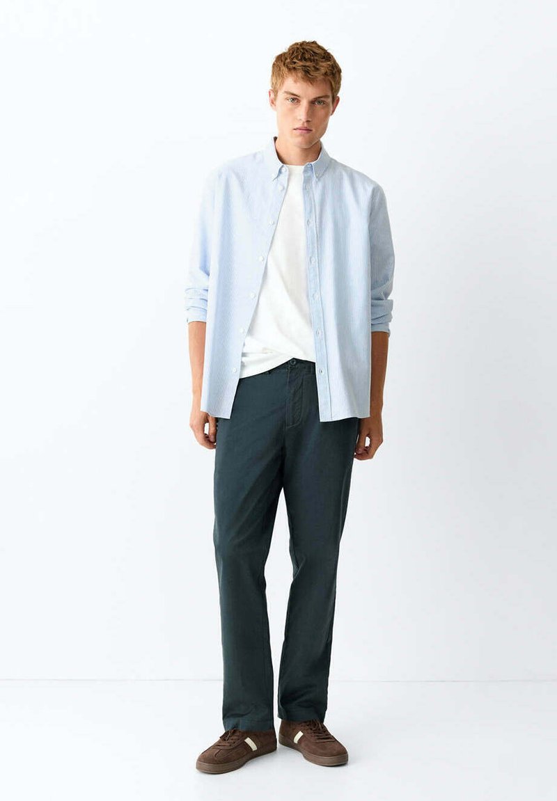 Jeune homme portant une chemise rayée bleu clair par-dessus un t-shirt blanc, un pantalon foncé et des chaussures décontractées marron, debout sur un fond blanc.