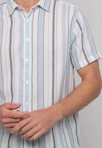 Chemise à manches courtes à boutons de couleur bleu clair, avec un motif rayé blanc et gris ; tissu texturé avec des coutures visibles ; col pointu.