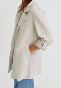 Femme portant un manteau beige clair à manches longues retroussées et un jean bleu, main dans la poche du manteau, sur un fond uni.