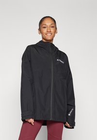 Crna vodootporna jakna s patentnim zatvaračem, podesivim kapuljačom, slobodnim krojem i Gore-Tex detaljima. Uparen s crvenim tajicama.