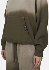 Hoodie vert olive avec un effet dégradé, comportant une poche avant et des poignets côtelés. Associé à un pantalon beige clair. Étiquette visible sur la poche.