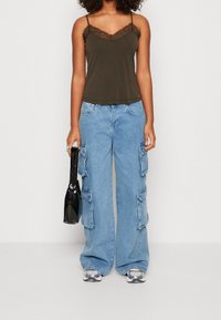 Olivgrön linne med spetskanter, tillsammans med ljusblå vida cargojackor med flera fickor. Hållare av en glänsande svart väska.