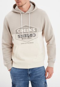 Sudadera con capucha en beige y crema con un bolsillo canguro frontal y estampado gráfico. Presenta cordones y puños acanalados para un ajuste adecuado.