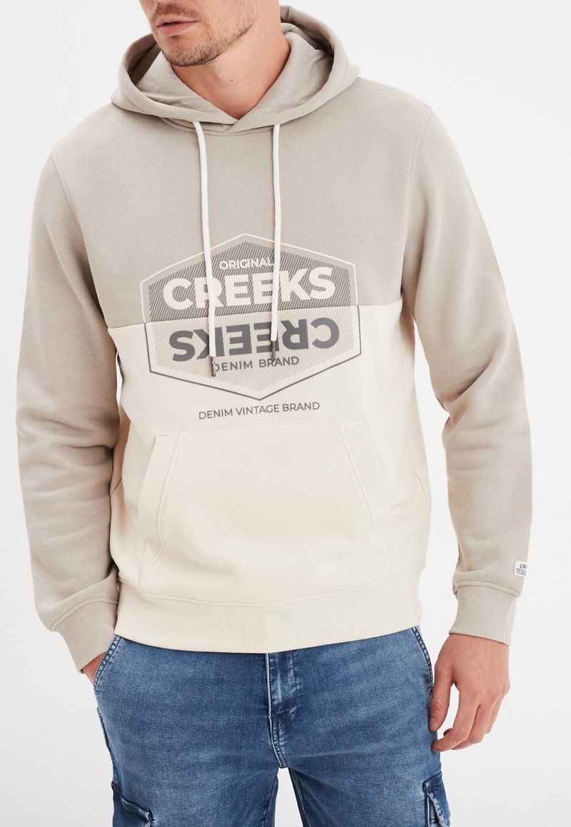 Sudadera con capucha en beige y crema con un bolsillo canguro frontal y estampado gráfico. Presenta cordones y puños acanalados para un ajuste adecuado.