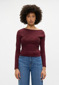 Gara piedurkne burgundijas krāsas blūze ar laivu formas kakla izgriezumu, rotāta ar izceltiem ziedu rakstiem, kas apvienota ar gaiši zila plaša iegriezuma džinsiem.