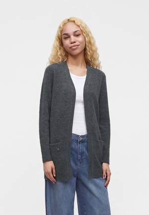 Jeune femme aux cheveux blonds bouclés porte un cardigan gris foncé ouvert devant, un haut blanc et un jean bleu ample sur un fond clair uni.