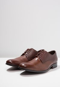 Chaussures en cuir marron avec une partie avant lisse et une zone de laçage texturée, dotées d'un bout arrondi et d'un petit talon. Design classique avec des accents minimaux.