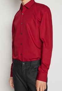 Rotes Langarmhemd mit einem Button-Down-Kragen, vorderen Knöpfen und schwarzen Akzenten. Wird mit schwarzer Hose getragen, die ein tailliertes Design aufweist.