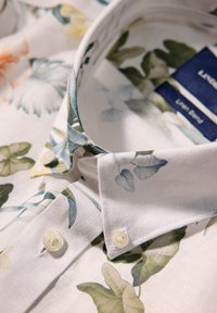 Camisa de mezcla de lino con un patrón floral en blanco, verde y amarillo suave. Presenta un cuello estructurado con dos botones.
