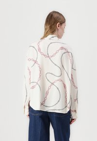 Blusa a maniche lunghe in tessuto leggero color crema con un motivo a catena in rosso e nero. Vestibilità comoda con orlo arrotondato e polsini con bottone.