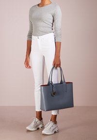 Szara skórzana torba na ramię z dwoma uchwytami, czerwoną podszewką i okrągłym dekoracyjnym charmsiem. Modelka nosi szary top, białe dżinsy i jasne trampki.