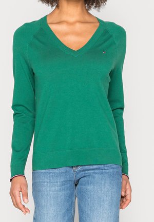 Frau trägt einen grünen Pullover mit V-Ausschnitt und langen Ärmeln sowie hellblaue Jeans, steht vor einem schlichten hellen Hintergrund.