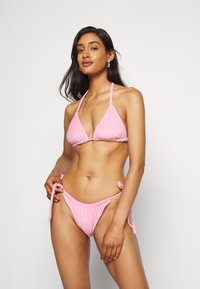 Conjunto de bikini em toalha de banho rosa, apresentando um top triangular com alças ajustáveis e uma parte de baixo com fechos laterais amarrados e detalhes franzidos.