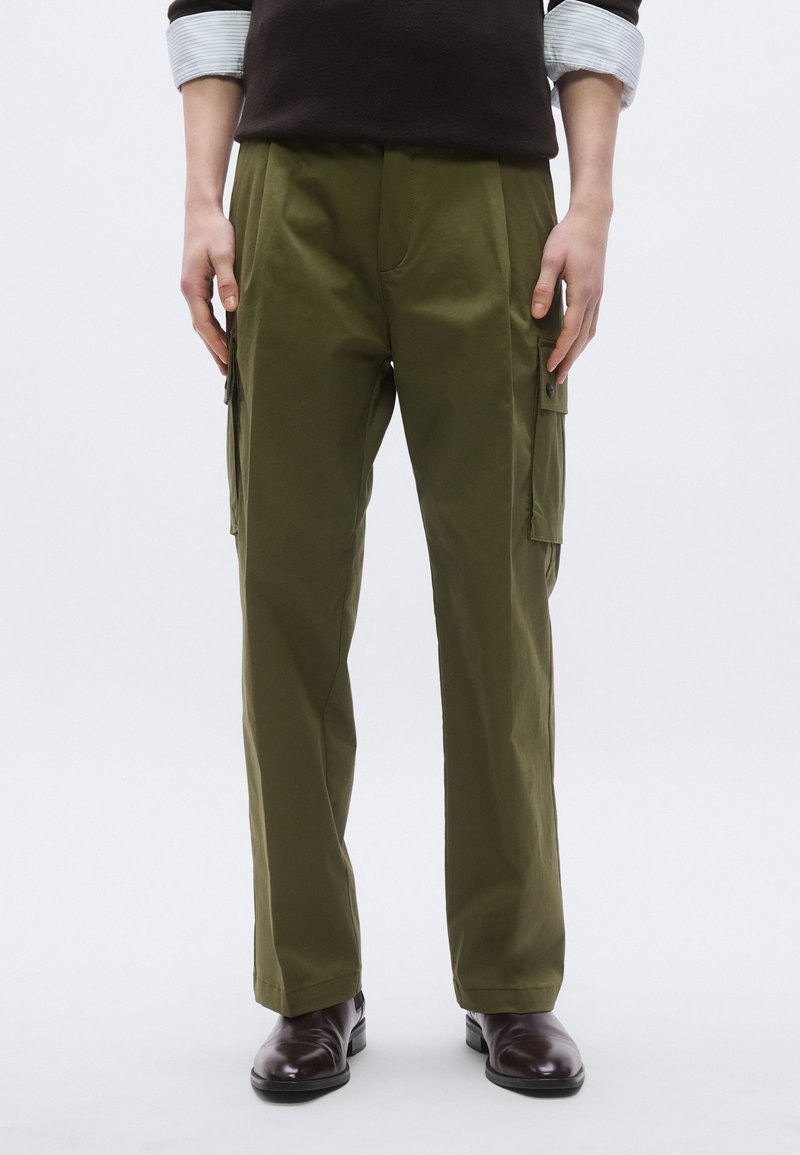 Olivzaļas kravas bikses ar sānu kabatām, izgatavotas no mīksta auduma, ar brīvu piegriezumu un redzamām šuvju detaļām. Uzvalkā profesionāla apavi.