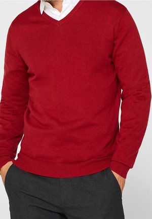 Man draagt een rode V-hals trui over een wit overhemd met kraag, met zijn handen in de zakken van een donkergrijze broek.