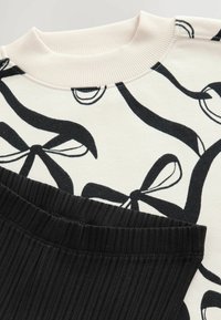 Bomullströja med svart bandmönster på krämfärgad bakgrund, med en ribbad halsringning. Svarta ribbade träningsbyxor med elastisk midja.