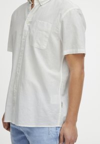 Camisa blanca de manga corta con botones, hecha de un tejido ligero y texturizado. Presenta un bolsillo de parche en el lado izquierdo del pecho, con un detalle de abertura lateral.
