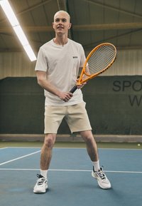 Tennisimängija valges, lühikeste varrukatega särgis, millel on "BOSS" logo, beežides lühikeses pükstes ja mustades-valgetes tennisetsippides, hoides oranži reketit.