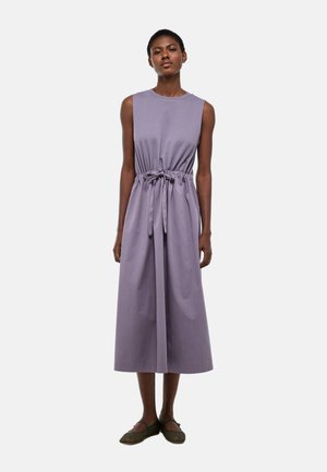 Femme debout, portant une robe midi lavande sans manches avec une ceinture nouée à la taille et des chaussures plates noires, sur fond blanc uni.