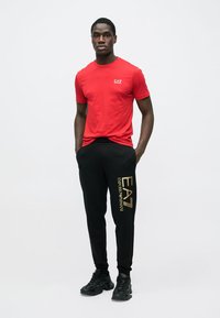 Červené bavlnené tričko s logom EA7 na hrudi, skombinované s čiernymi teplákmi s nápisom "EA7 Emporio Armani" v zlatej farbe. Čierne tenisky.