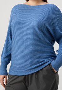 Maglione blu a coste con scollatura ampia, maniche lunghe e vestibilità comoda. Il tessuto testurizzato contrasta con i pantaloni grigio scuro lisci.