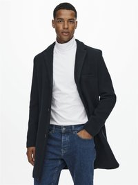 Manteau en mélange de laine navy avec des revers crantés, poche poitrine unique et fermeture à boutons sur le devant, superposé à un col roulé blanc et un jean bleu.