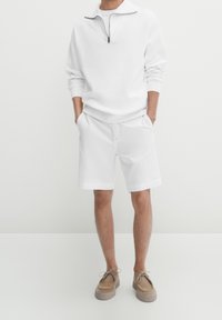 Uomo che indossa una felpa bianca a maniche lunghe con zip a quarto, pantaloni corti bianchi fino al ginocchio e scarpe casual beige con lacci, in piedi con le mani in tasca.