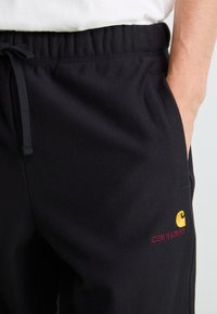 Pantaloni de trening negri, din material moale, cu talie elastică, având un buzunar lateral și un logo galben brodat cu text roșu.