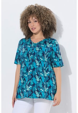 Kvinde med krøllet hår iført en kortærmet skjorte med blå tropiske blade og fuglemønster samt hvide bukser, stående foran en hvid væg.