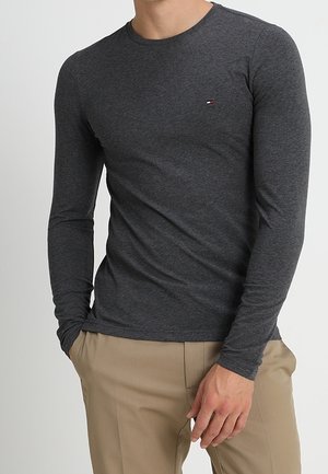 Langärmeliges Rundhals-Shirt aus dunkelgrauem Baumwollmischgewebe mit unauffälligem Logo auf der Brust und glatter Textur. Kombiniert mit beigen Hosen.