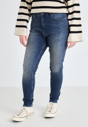 Skinny blue jeans with a faded finish, high waist, and five-pocket design, paired with a striped long-sleeve sweater and cream sneakers.