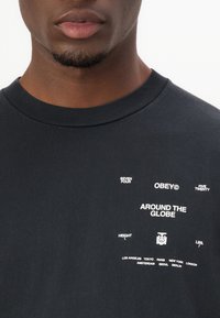 Black cotton t-shirt with crew neck. Features white text including “OBEY,” “AROUND THE GLOBE,” and city names in various fonts.