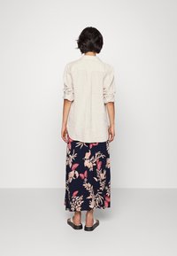 Person med kort mörkt hår, iklädd en ljusbeige långärmad skjorta, marinblå kjol med rosa blommor och fjärilstryck, samt vita sandaler.