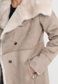 Beige kabát s umělou kožešinou, dvojité zapínání, stříbrné knoflíky a boční kapsy. Měkká textura s hladkým povrchem.