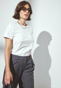 Valkoinen lyhythihainen t-paita yhdistettynä harmaisiin housuihin. T-paidassa on sileä pinta, ja housuissa on klassinen malli, jossa on etuosan laskokset.