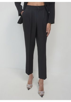 Kobieta ubrana w czarne, dopasowane spodnie, czarną marynarkę i srebrne, spiczaste szpilki z ozdobami, stojąca na tle jednolitego jasnego tła.