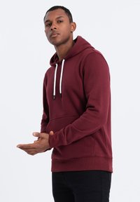 Felpa bordeaux con tasca frontale, cappuccio con coulisse e punte nere, e polsini a costine. Il materiale sembra morbido con una texture liscia.