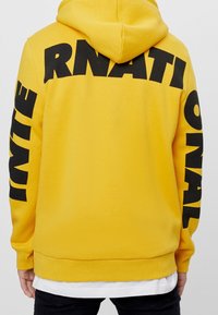 Żółta bluza z kapturem z dużym czarnym napisem "INTERNATIONAL" na plecach oraz czarnym tekstem na rękawach. Materiał mieszanki bawełny, luźny krój.