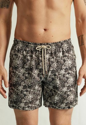 Bañador corto de hombre con estampado floral en blanco y negro, cintura con cordón y clip lateral, llevado por una persona sin camisa con tatuajes.