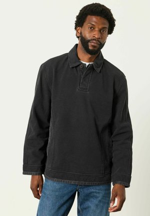 Man with short curly hair and beard wearing a black long-sleeve pullover and blue jeans standing against a plain light background.