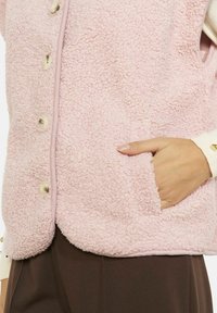 Fliisa vestēra gaiši rozā krāsā ar tekstūru; tam ir sprādzīšu aizdare un sānu kabata. Apakšmala ir pabeigta ar mīkstu apdari.