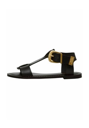 Sandalo piatto nero con una fascia larga, hardware in oro e design minimalista. Presenta una finitura liscia e cuciture a contrasto.