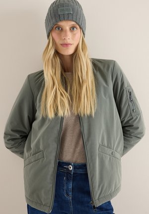 Olivově zelená bomber bunda s vroubkovanými manžetami, bočními kapsami a zipem. V kombinaci se šedou vroubkovanou čepicí. Modelka má dlouhé blond vlasy.