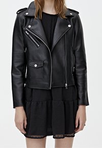 Femme portant une veste de motard en cuir noir avec des fermetures éclair argentées sur une robe noire texturée, debout devant un fond clair uni.