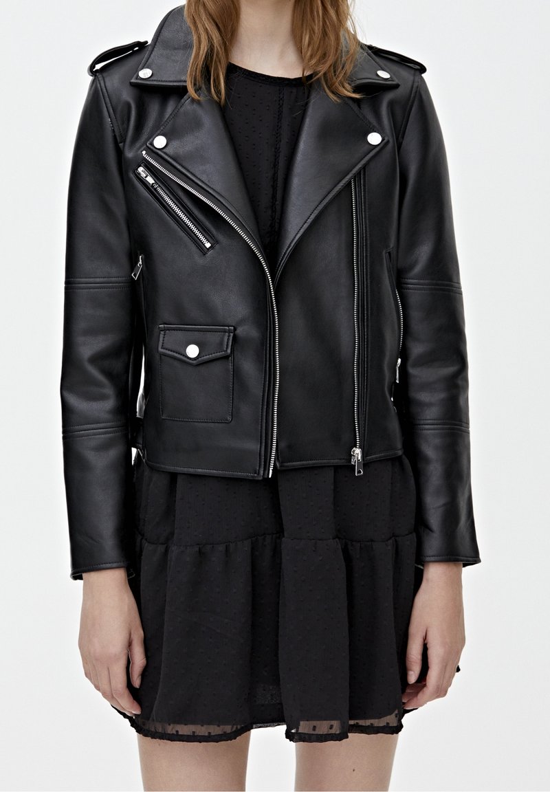 Femme portant une veste de motard en cuir noir avec des fermetures éclair argentées sur une robe noire texturée, debout devant un fond clair uni.