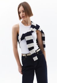 Vit linne tillsammans med mörka, högmidjade jeans. En randig marinblå och krämfärgad tröja hänger avslappnat över axlarna. Guldbälte som detalj.