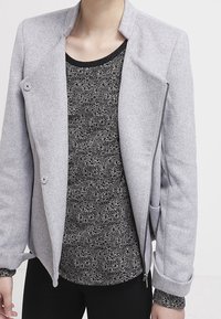 Chaqueta gris, estructurada, con un alto cuello, dos grandes botones y un zipper asimétrico. Camiseta interna con un patrón en blanco y negro visible debajo.