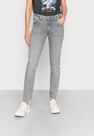 Henkilö pukeutunut slim fit -mallisiin vaaleanharmaisiin farkkuihin ja valkoisiin tennareihin, toisessa kädessä taskussa ja musta graafinen t-paita.