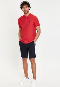 Camiseta polo roja de manga corta, con un sutil logo en el pecho, combinada con pantalones cortos azul marino oscuros y zapatillas blancas.